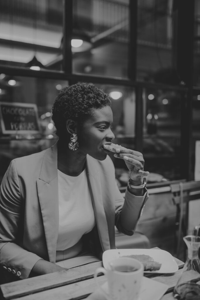 African American elegant woman eating sandwich and sitting at table with cup of drink in street cafe